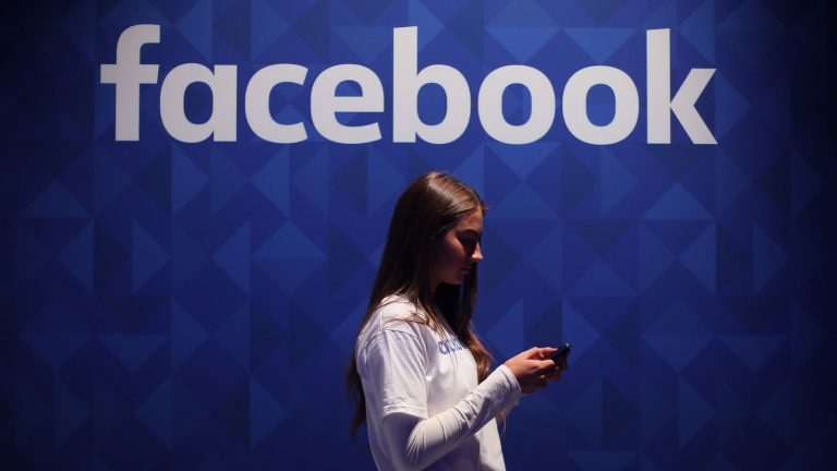 Junge Frau blickt auf Smartphone vor Wand mit Facebook-Schriftzug