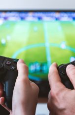 Nahaufnahme der Hände von zwei Personen, die jeweils einen Controller halten. Auf dem Monitor ist ein Fußballspiel zu erkennen.