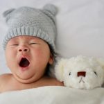 Gähnendes Baby mit Strickmütze und Teddy im Bett