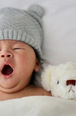 Einschlafhilfe für Babys und Erwachsene: So funktionieren elektronische Helfer Gähnendes Baby mit Strickmütze und Teddy im Bett