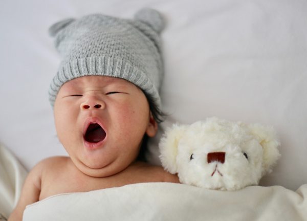 Gähnendes Baby mit Strickmütze und Teddy im Bett
