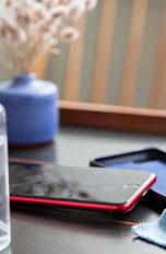 Ein iPhone SE in roter Farbe liegt auf dem Tisch. Daneben sind eine Schutzhülle, eine Sprühflasche und ein Mikrofasertuch zu sehen.