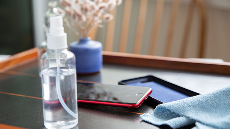 Smartphone desinfizieren Ein iPhone SE in roter Farbe liegt auf dem Tisch. Daneben sind eine Schutzhülle, eine Sprühflasche und ein Mikrofasertuch zu sehen.