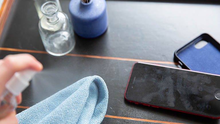 Smartphone desinfizieren Eine Person sprüht etwas Hygienemittel auf ein Mikrofasertuch. Daneben liegen das verschmutzte Smartphone und eine Hülle.