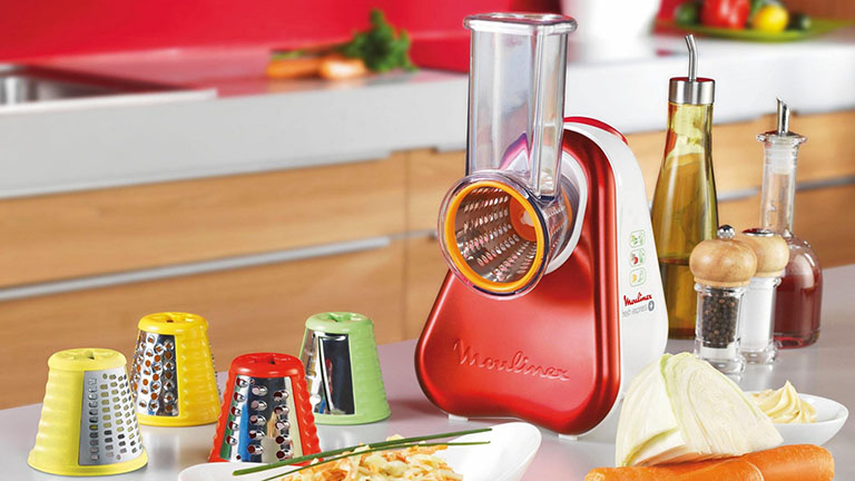 Moulinex Zerkleinerer Der Zerkleiner von Moulinex steht zusammen mit mehreren Aufsätzen und verarbeitetem Gemüse auf einem Küchentisch.