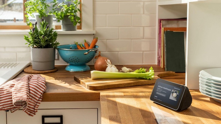 Amazon Echo Show 5 Das Amazon Echo Show steht in einer Küche. Im Hintergrund ist ein Schneidebrett mit Gemüse zu sehen. Das Gerät zeigt ein Rezept für Hühner-Nudelsuppe an.