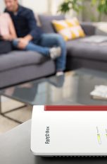 Eine FritzBox auf einem Sideboard. Im Hintergrund sind zwei Personen vor einem Laptop zu sehen.