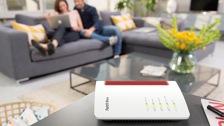 Eine FritzBox auf einem Sideboard. Im Hintergrund sind zwei Personen vor einem Laptop zu sehen.