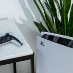 Die PlayStation 5 steht vor einer Pflanze. Daneben liegt der DualSense-Controller erhöht auf einem Tisch.