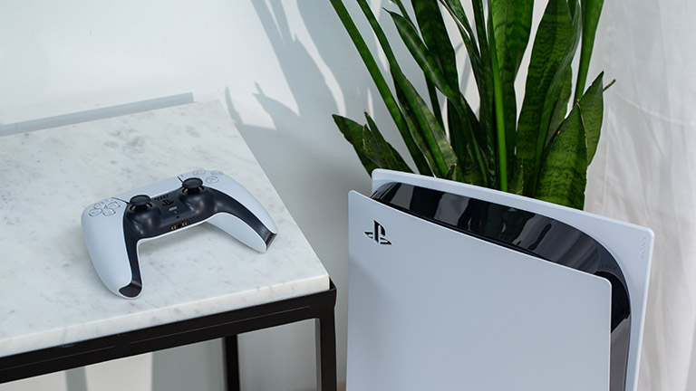 Die PlayStation 5 steht vor einer Pflanze. Daneben liegt der DualSense-Controller erhöht auf einem Tisch.