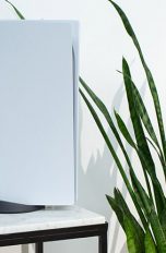Die PlayStation 5 steht dekorativ auf einem Marmortisch neben einer Zimmerpflanze. An die Konsole angelehnt steht der neue DualSense-Controller.