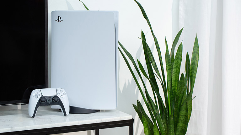 Die PlayStation 5 steht dekorativ auf einem Marmortisch neben einer Zimmerpflanze. An die Konsole angelehnt steht der neue DualSense-Controller.