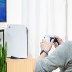 Eine Person sitzt vor einer PlayStation 5 und hält dabei den neuen DualSense-Controller in der Hand.