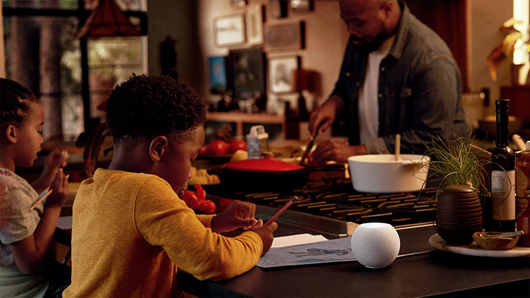 HomePod Zwei Kinder und eine Person sind in der Küche. Auf dem Tisch steht ein HomePod mini in Weiß.