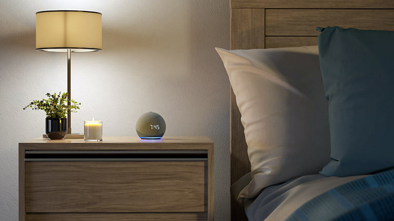 Amazon Echo Dot Ein Echo Dot mit Uhr-Anzeige und leuchtendem Ring neben dem Bett.