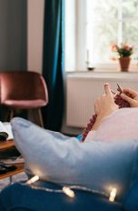 Eine Person sitzt auf dem Sofa und strickt. Nebenbei sieht sie fern.