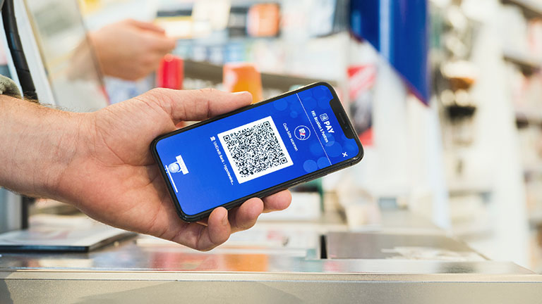 Payback Pay Eine Person hält ein Smartphone mit QR-Code von Payback Pay in Richtung einer Kasse.