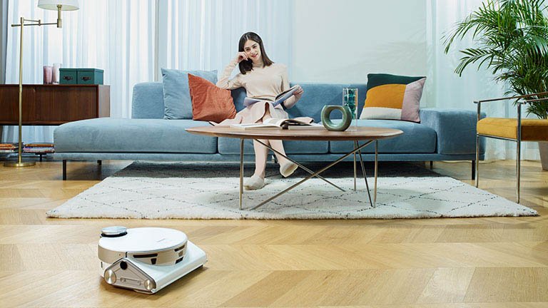 Samsung JetBot 90 AI Der JetBot 90 AI+ fährt durch ein Wohnzimmer. Im Hintergrund sitzt eine Person auf einem Sofa.