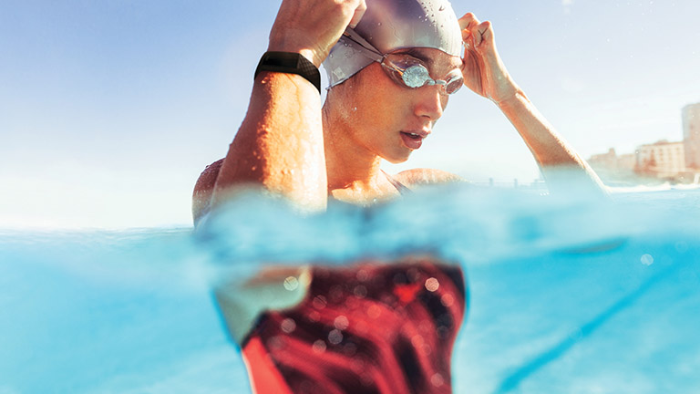 Fitbit Charge 4 Eine Schwimmerin im Wasser setzt sich ihre Schwimmbrille auf. Am Handgelenk trägt sie das wasserabweisende Fitness-Armband Fitbit Charge 4.