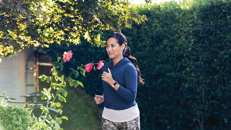 Fitbit Inspire 2 Eine Frau joggt durch eine gartenähnliche Kulisse. Sie trägt einen blauen Kapuzenpullover und am Handgelenk das Fitness-Armband Inspire 2 von Fitbit.
