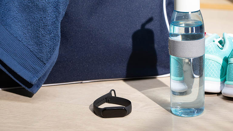 Xiaomi MI Smart Band 5 Das Fitness-Armband Mi Smart Band 5 von Xiaomi liegt vor einer großen, blauen Sporttasche. Daneben befinden sich eine Wasserflasche und ein Paar Sportschuhe.