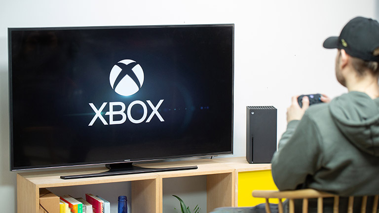 Eine Person sitzt vor einer Xbox Series X. Auf dem Fernseher ist der Startbildschirm zu sehen.