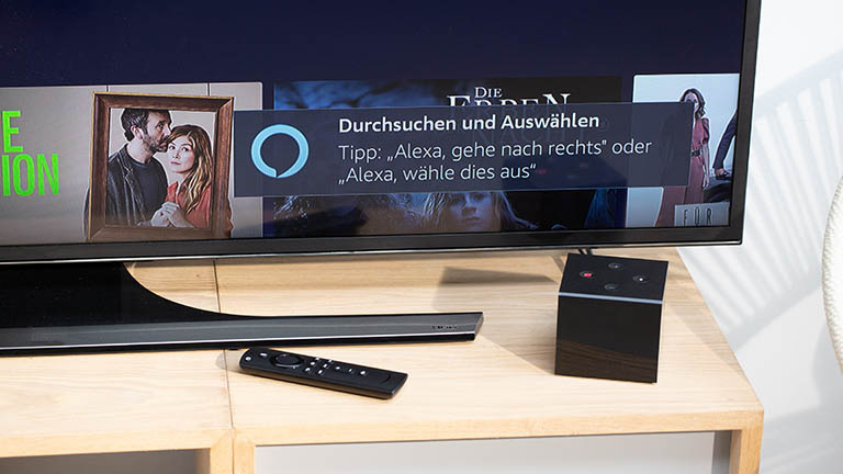 Amazon Fire TV Cube Ein TV-Gerät, auf dem ein Tipp für die Steuerung per Alexa angezeigt ist. Vor dem Fernseher steht der Fire TV Cube.