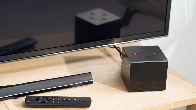 Der Amazon Fire TV Cube steht vor einem Fernseher. Daneben ist die Fernbedienung zu sehen.