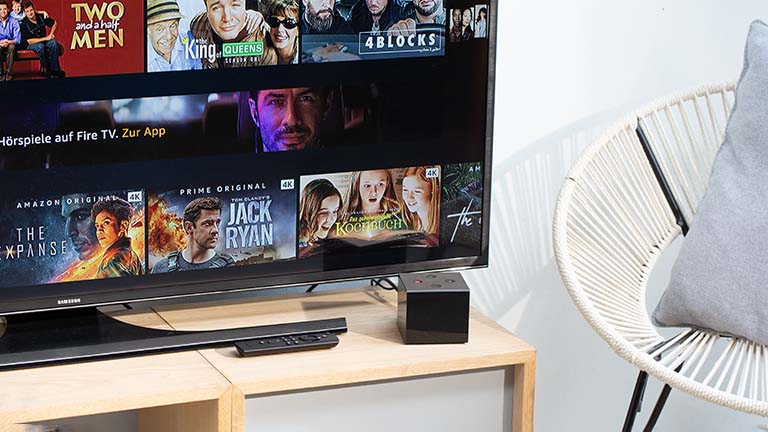Amazon Fire TV Cube Der Fire TV Cube von Amazon vor einem Fernseher. Auf dem Bildschirm ist das Hauptmenü zu sehen.