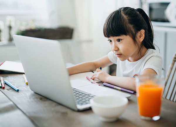 Ein junges Mädchen sitzt an einem Küchentisch vor einem aufgeklappten Laptop. Neben ihr befinden sich ein paar Stifte und ein volles Glas Saft.