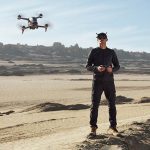 Ein Mann steht in sandiger Umgebung. Er hält einen Drohnencontroller in der Hand und trägt die FPV-Brille von DJI. Vor ihm fliegt die DJI FPV.