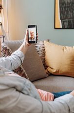 Eine Person sitzt auf dem Sofa und telefoniert per Videoübertragung mit einer anderen Person.