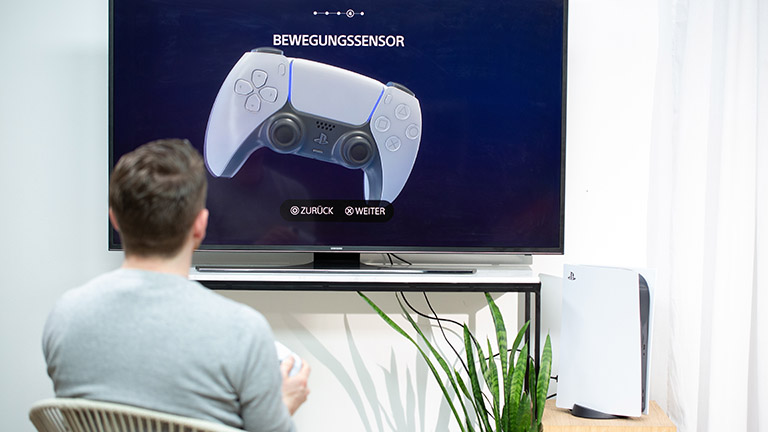 PS 5 einrichten Eine Person sitzt vor einem Fernseher. Auf diesem ist der Einrichtungsprozess der PS5 zu sehen. Die Konsole selbst steht neben dem TV.