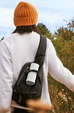 Ein Mann streift durch ein Feld. An seinem Rucksack hängt der Bluetooth-Lautsprecher Sonos Roam in weiß.