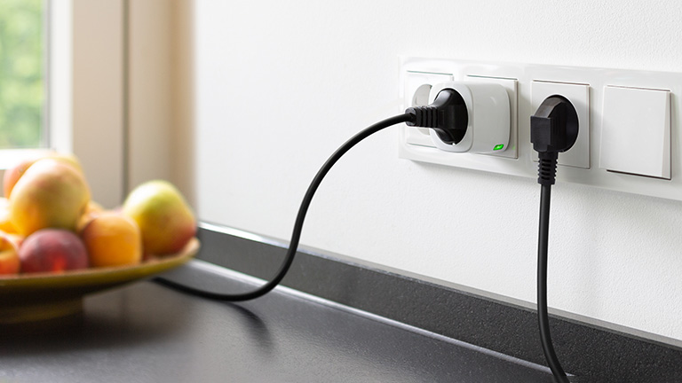 Eve Energy Eine smarte Eve Energy steckt in einer Wand. Angeschlossen ist ein Gerät mit schwarzem Kabel.