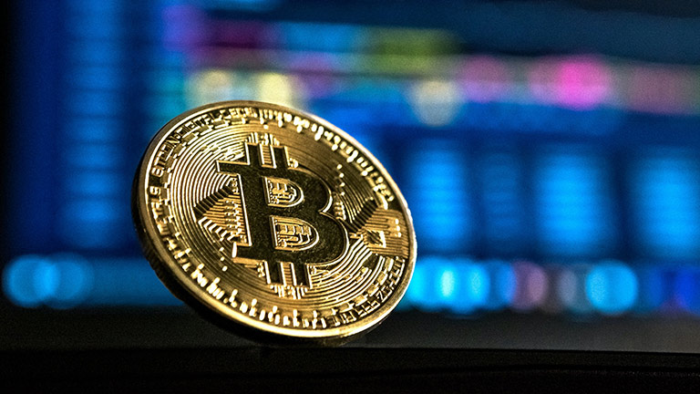 Bitcoin Aufnahme einer Goldmünze mit dem Bitcoin-Logo, im unscharfen Hintergrund befindet sich ein Bildschirm.