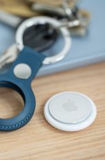 Ein Apple AirTag liegt neben einem Schlüsselanhänger. Im Hintergrund sind ein paar Euro-Münzen für den Größenvergleich zu sehen.)