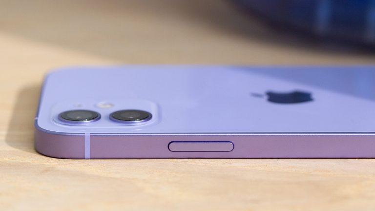 iPhone 12 Das iPhone 12 liegt bäuchlings auf einem Holztisch. Zu erkennen ist die Einschalttaste im Rahmen des Smartphones und die Dualkamera auf der Rückseite.