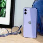 Das iPhone 12 in der neuen Farbe Violett steht auf einem Holztisch. Daneben liegt eine farblich passende Brille.