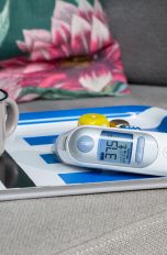 Auf einem Tablett steht eine Emailletasse mit Tee, daneben liegt ein Infrarotfieberthermometer.