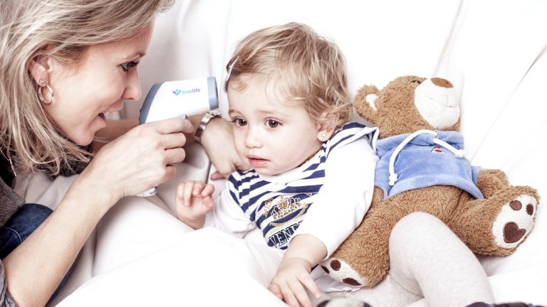 TrueLife Care Q7 Eine Mutter misst die Temperatur bei ihrem Kind, das mit einem Kuscheltier im Bett liegt.