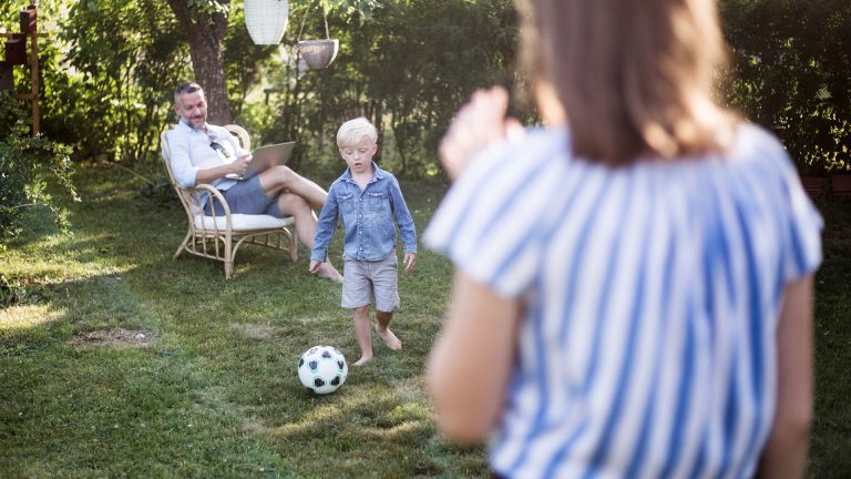 Eine Person sitzt auf einem Stuhl im Garten, während ihr Sohn Anlauf nimmt, um einen Ball zu schießen, eine weitere Person steht im Vordergrund und schaut zu.