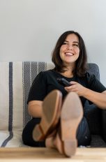 Eine Frau sitzt auf dem Sofa, legt die Beine entspannt auf dem Tisch ab und hält in der Hand eine Fernbedienung.