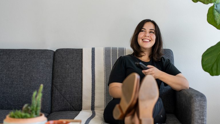 Eine Frau sitzt auf dem Sofa, legt die Beine entspannt auf dem Tisch ab und hält in der Hand eine Fernbedienung.