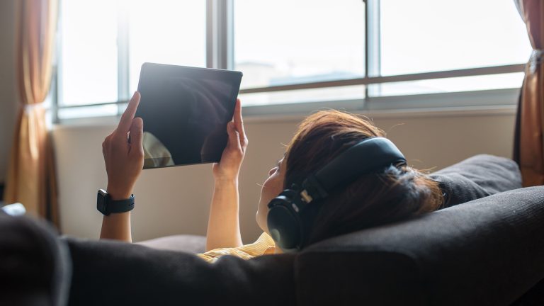 Netflix Eine Frau liegt auf dem Sofa, in der Hand ein Tablet, auf dem Kopf einen Kopfhörer.