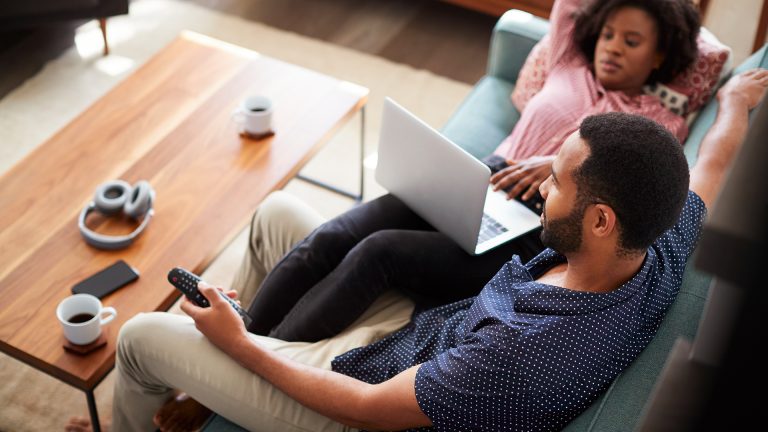 Netflix Ein Paar sitzt auf dem Sofa, ihre Beine hat sie über seine Oberschenkel gelegt, auf ihrem Schoß ein Laptop, er schaut zum Fernseher, in der Hand eine Fernbedienung.