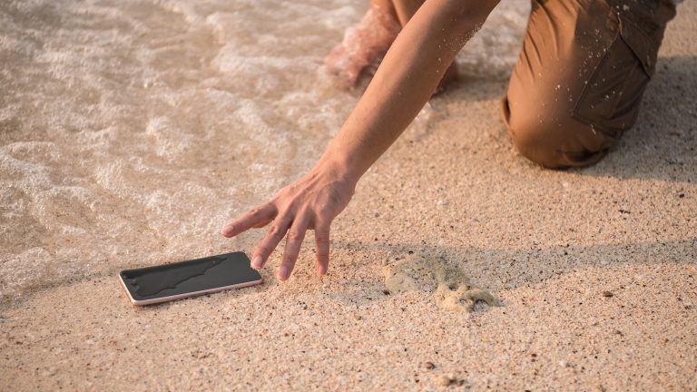 Smartphone ins Wasser gefallen Ein Handy liegt am Strand und wird von Wasser umspült, eine Hand greift nach dem Gerät.