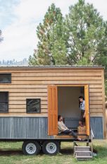 Zwei Personen sitzen am Eingang eines Tiny House, das auf einen Anhänger gebaut ist.