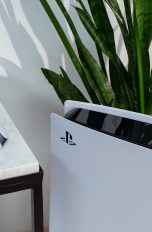 Die PlayStation 5 steht vor einer Dekopflanze. Daneben liegt der DualSense-Controller erhöht auf einem Tisch.
