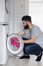 In einem Badezimmer befüllt ein Mann die Waschmaschine, über der Waschmaschine steht ein Wäschetrockner
